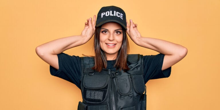 A female police officer in a black uniform and cap smiles while holding her hands above her head.