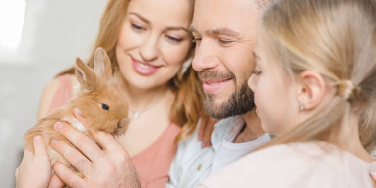 A family of three, including a child, smiles while holding a small brown rabbit indoors.